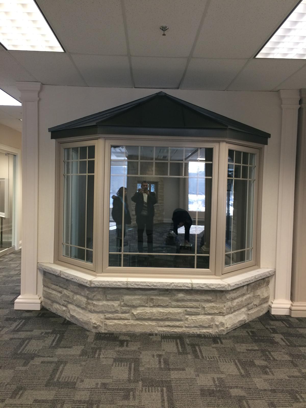 Three-panel bay window in beige vinyl with grid pattern, set on a stone base, indoor showroom.