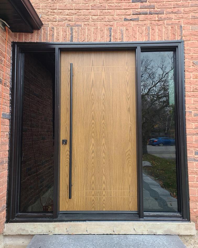 Exterior view of a modern oak-finish fiberglass entry door with black frame and tinted sidelites against a brick wall.